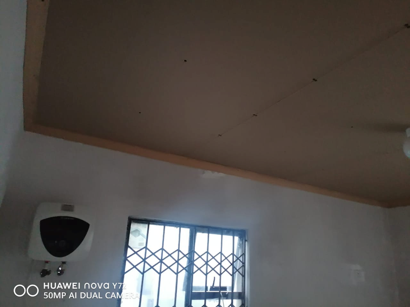 An interior space featuring an unfinished ceiling, a wall-mounted electric water heater, and a window fitted with a security gate.
