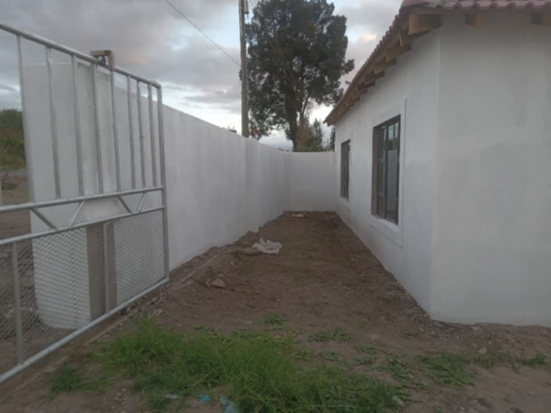 Exterior view of the property showing a perimeter wall, metal gate, and unfinished ground area surrounding the building structure.
