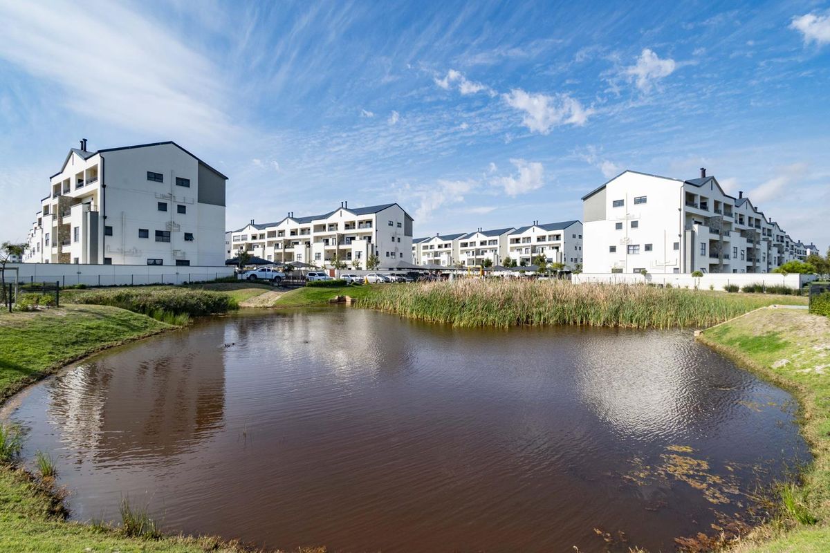 A scenic view of a modern apartment complex featuring multi-story residential buildings overlooking a landscaped pond and green space.