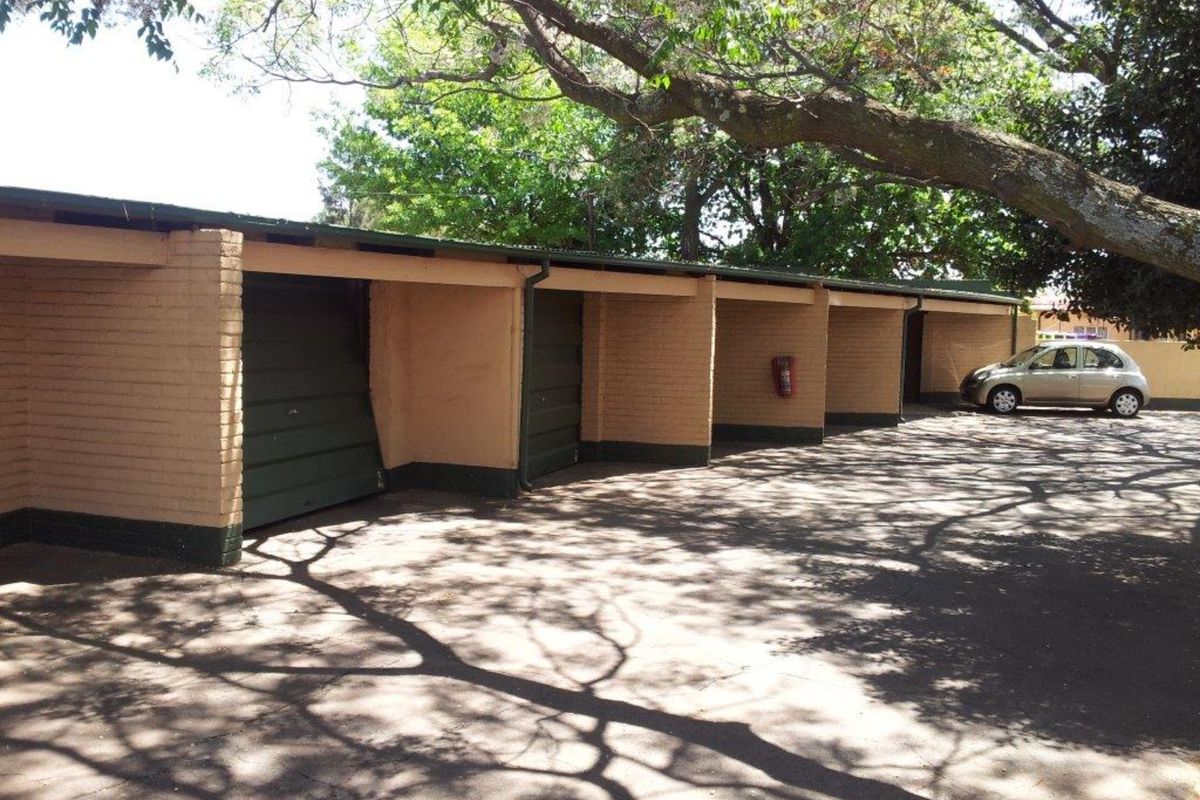 A row of open-style brick garages with a damaged door, situated under mature trees with an asphalt driveway.
