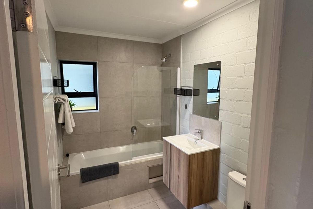 Modern bathroom featuring a bathtub with glass shower screen, wall-mounted vanity with basin, and a toilet, finished with neutral floor and wall tiling.