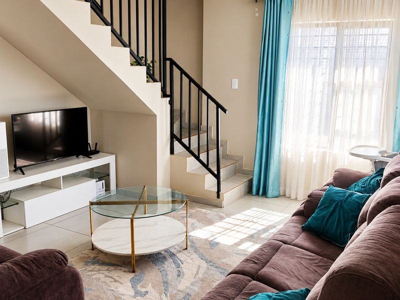 A bright living room featuring a staircase with a metal balustrade, tiled flooring, and natural light from a large window.