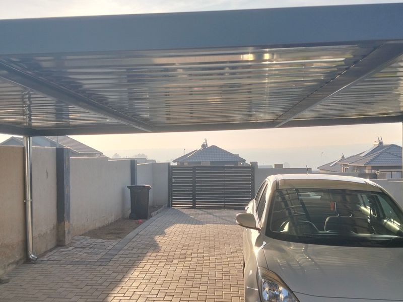 A covered carport area featuring a metal roof structure, paved driveway, and secure boundary wall with a metal gate.