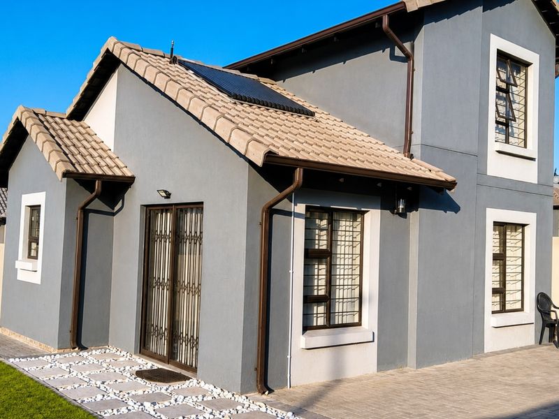 A well-maintained rear exterior view of a modern house featuring a tiled roof, solar water heating system, paved patio area, and secure window bars.