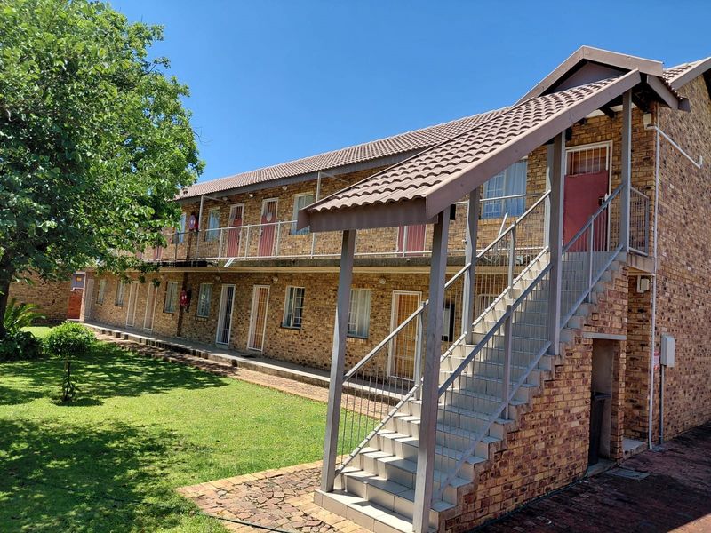 Exterior view of a multi-story face brick apartment complex featuring an external staircase, metal balustrades, and a well-maintained communal garden area.