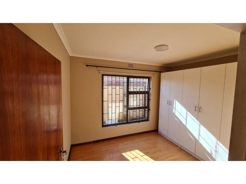 A bright, empty bedroom featuring laminate flooring, built-in cupboards, and a window fitted with security bars.