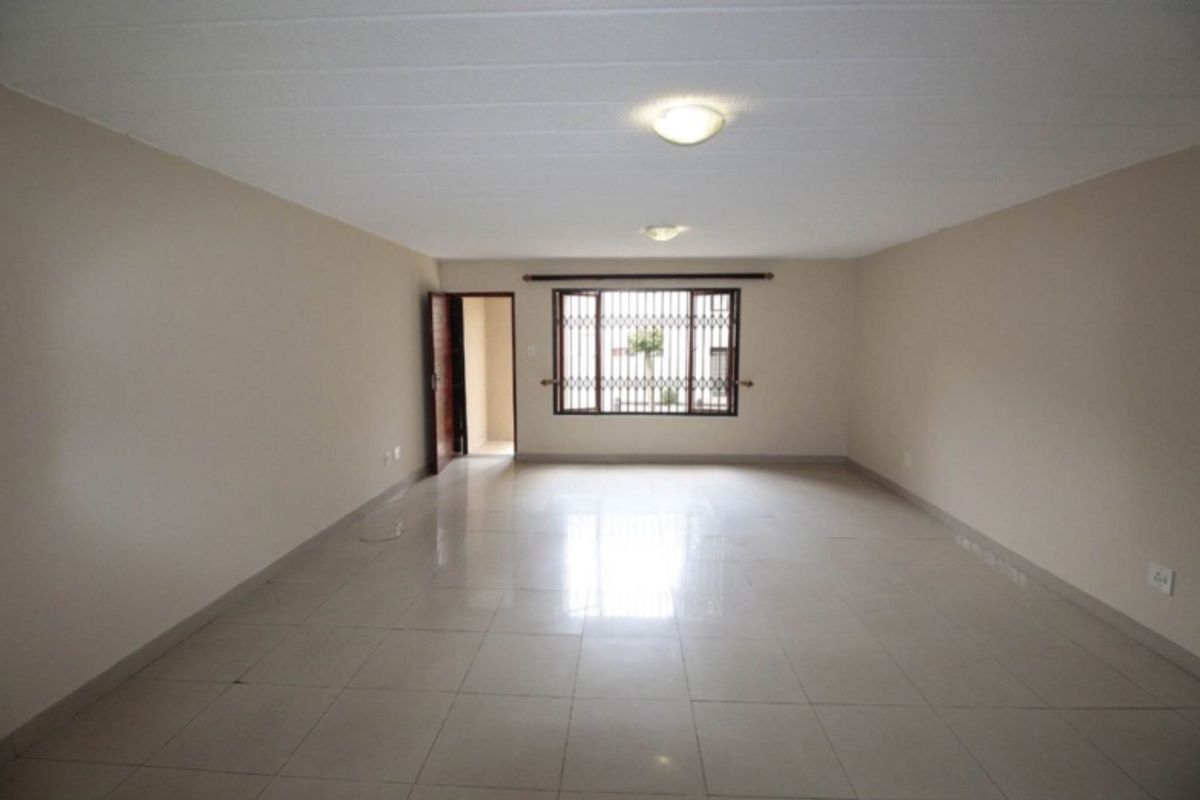 A spacious, empty living area featuring neutral tiled flooring, plain walls, and a window fitted with security bars.
