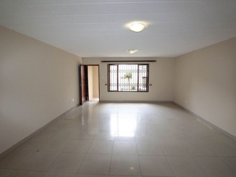 A spacious, empty living area featuring neutral tiled flooring, plain walls, and a window fitted with security bars.