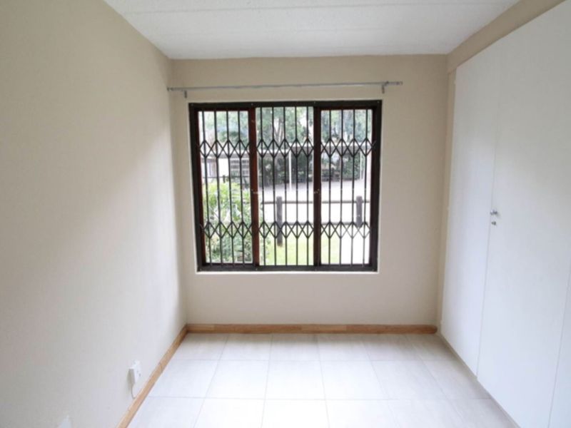 A bright, empty bedroom featuring tiled flooring, built-in cupboards, and a window fitted with security bars.