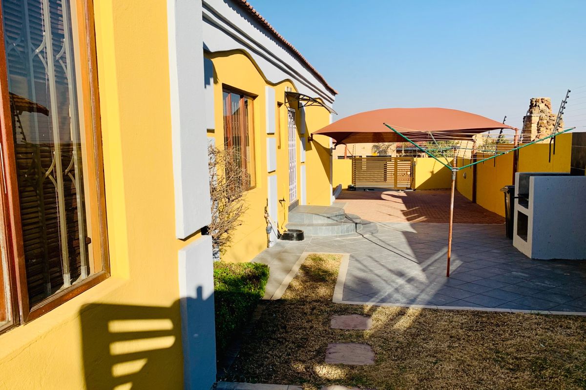 A view of the rear exterior of the cottage, featuring a paved patio area, a built-in braai, and a shade sail carport.