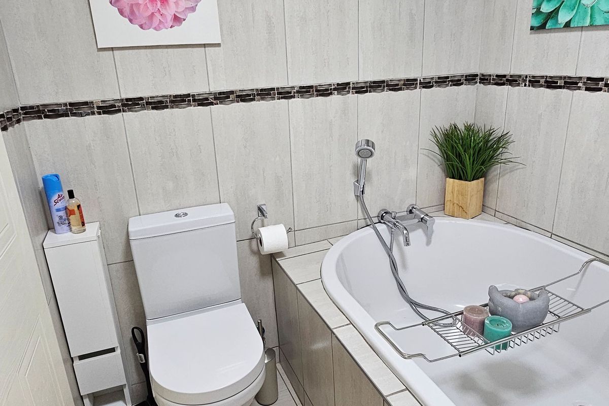 A well-maintained bathroom featuring a built-in bathtub with a handheld shower mixer, a standard toilet, and neutral floor-to-ceiling tiling with a decorative accent border.