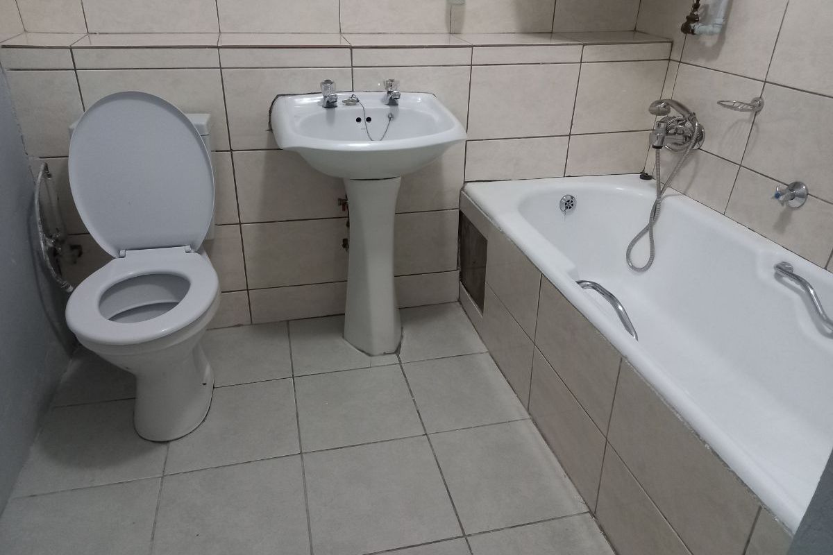 A functional bathroom featuring a pedestal basin, toilet, and bathtub with a handheld shower mixer, finished with neutral ceramic wall and floor tiles.