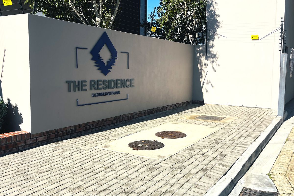 The entrance facade of The Residence in Bloubergstrand, featuring a paved driveway, boundary wall with signage, and integrated security fencing.