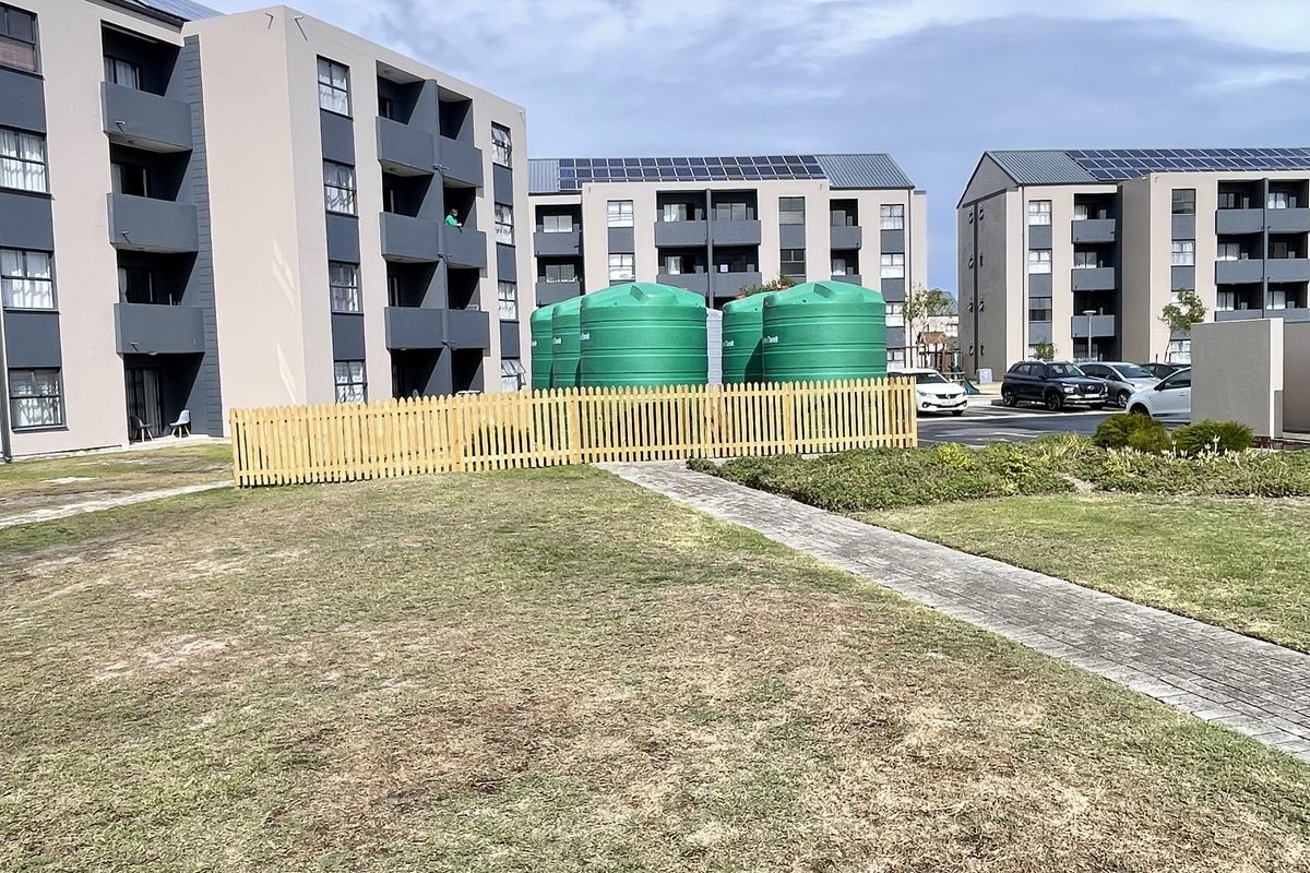 Exterior view of apartment complex buildings featuring solar panels, water storage tanks, and landscaped grounds with a paved walkway.