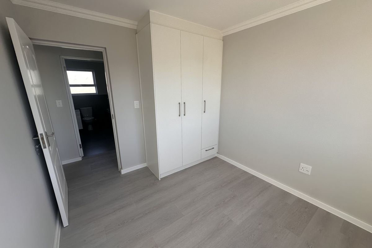 A bright, neutral-toned bedroom featuring modern laminate flooring, built-in white cabinetry, and direct access to an en-suite bathroom.