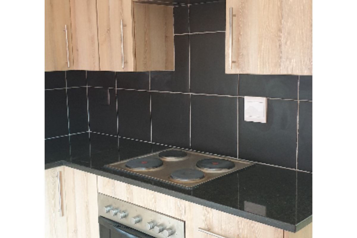 A compact kitchen area featuring light wood-finish cabinetry, black granite countertops, a built-in electric hob, and an integrated oven with a dark tiled backsplash.