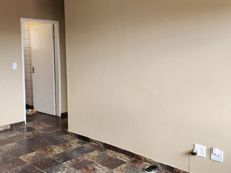 An empty open-plan living area featuring tiled flooring, built-in shelving, and visible kitchen plumbing connections, leading to an adjacent room.