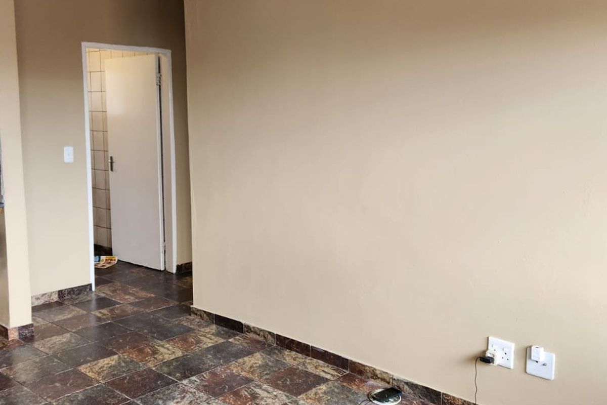 An empty open-plan living area featuring tiled flooring, built-in shelving, and visible kitchen plumbing connections, leading to an adjacent room.