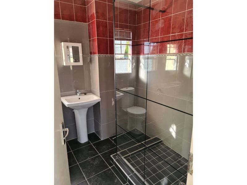 A functional bathroom featuring a pedestal basin, a glass-enclosed shower, and a toilet, finished with contrasting red and grey wall tiles.