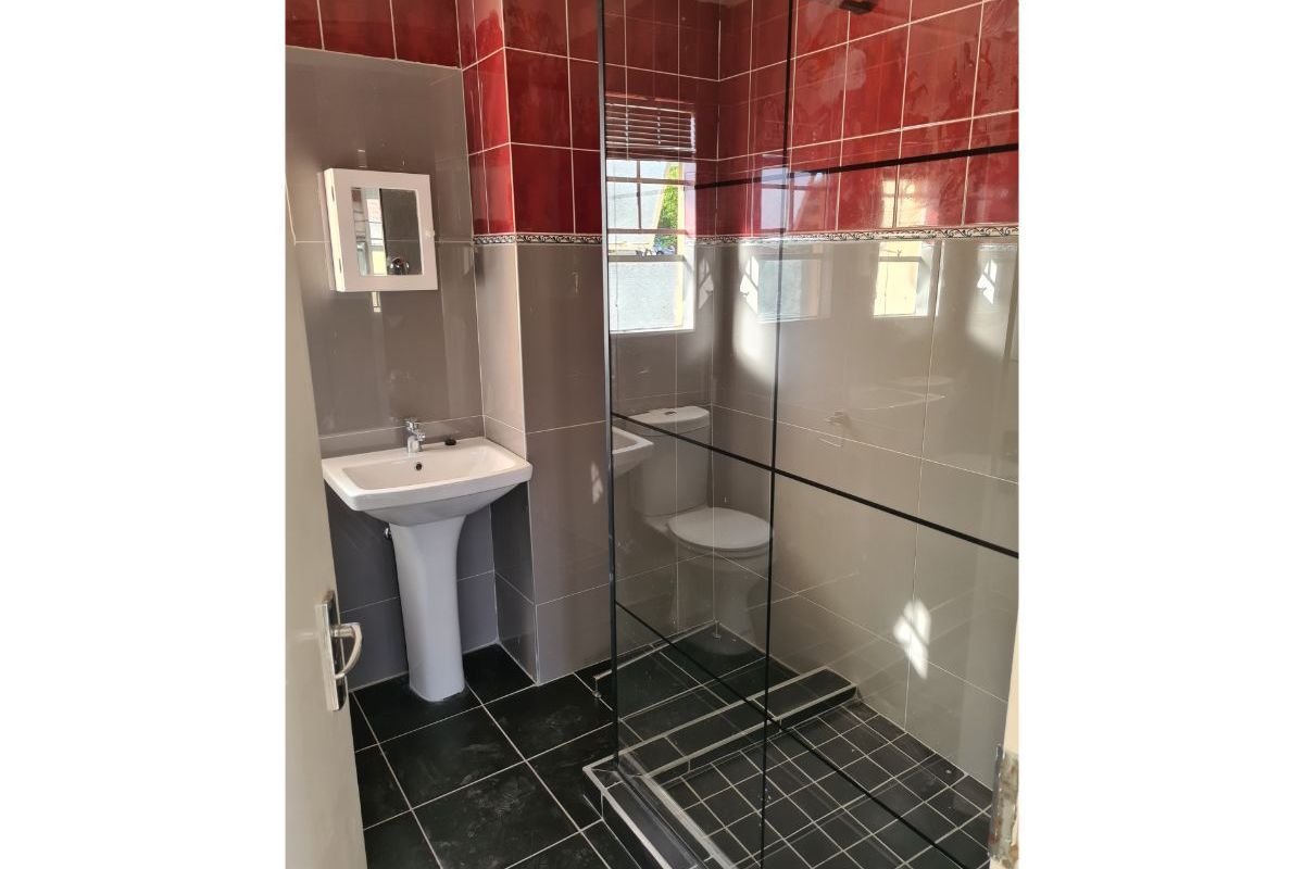 A functional bathroom featuring a pedestal basin, a glass-enclosed shower, and a toilet, finished with contrasting red and grey wall tiles.