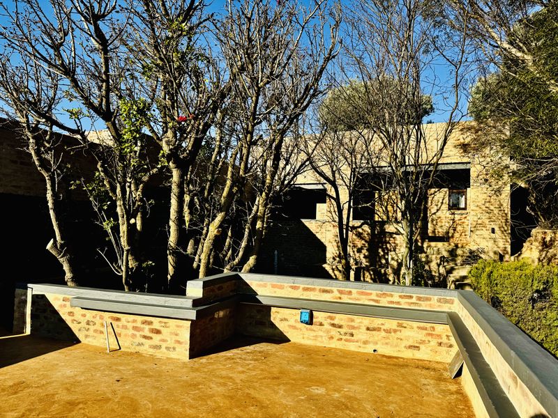 An outdoor patio area featuring built-in brick seating and a concrete floor, set against a brick building facade with mature trees.