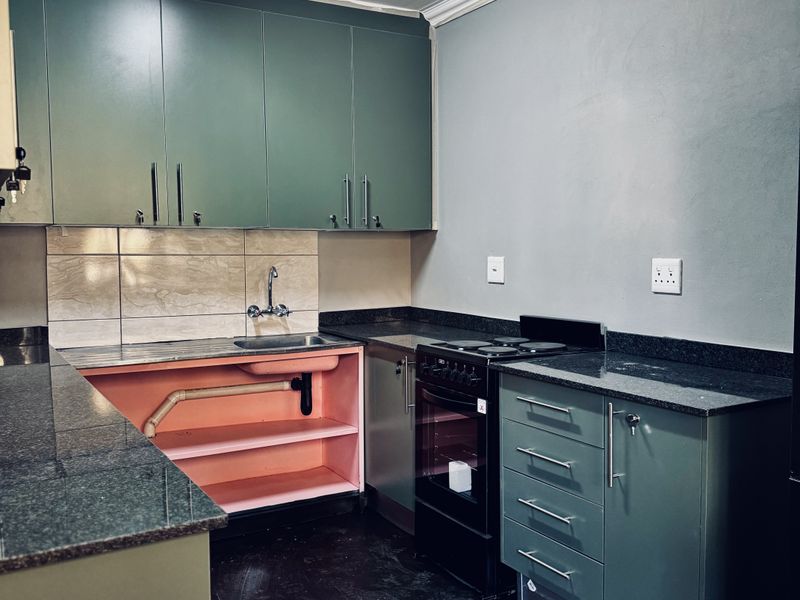 A functional kitchen featuring green cabinetry, granite countertops, and a tiled backsplash, currently requiring repairs to the under-sink cabinetry and flooring.