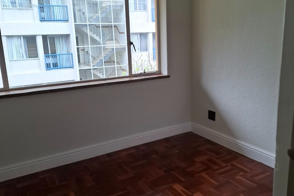 An empty bedroom featuring well-maintained parquet flooring, neutral walls, and a large window providing natural light and an urban view.