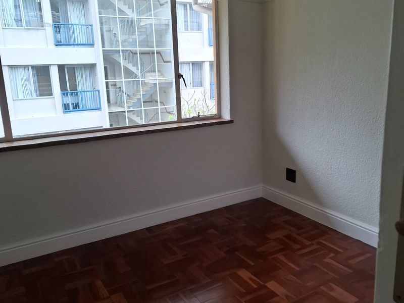 An empty bedroom featuring well-maintained parquet flooring, neutral walls, and a large window providing natural light and an urban view.