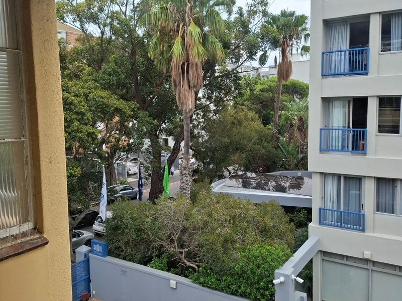 An elevated view overlooking the apartment complex grounds, featuring paved driveway areas, mature trees, and the adjacent building facade with blue-railed balconies.