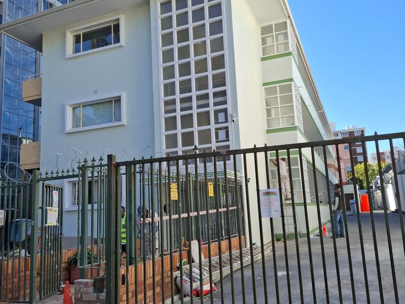 Exterior view of an apartment building facade featuring a metal security gate, brick boundary wall, and distinctive grid-pattern window design.