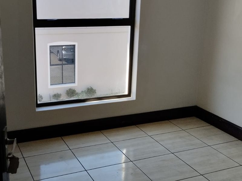 A bright, empty bedroom featuring neutral tiled flooring, dark skirting boards, and a large window with an aluminium frame.