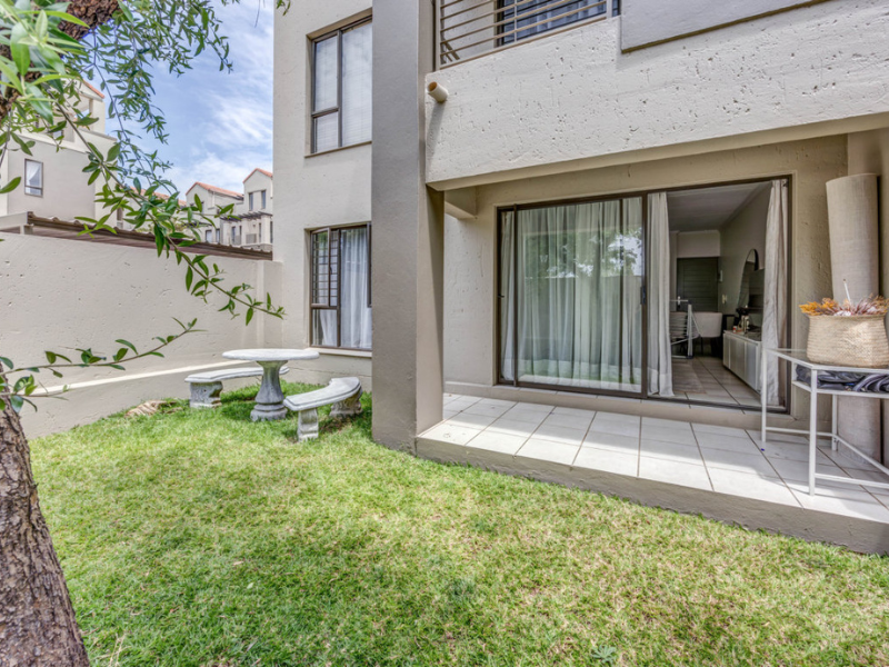 A private rear garden featuring a tiled patio area, accessible via sliding glass doors, set against the apartment building's exterior wall.