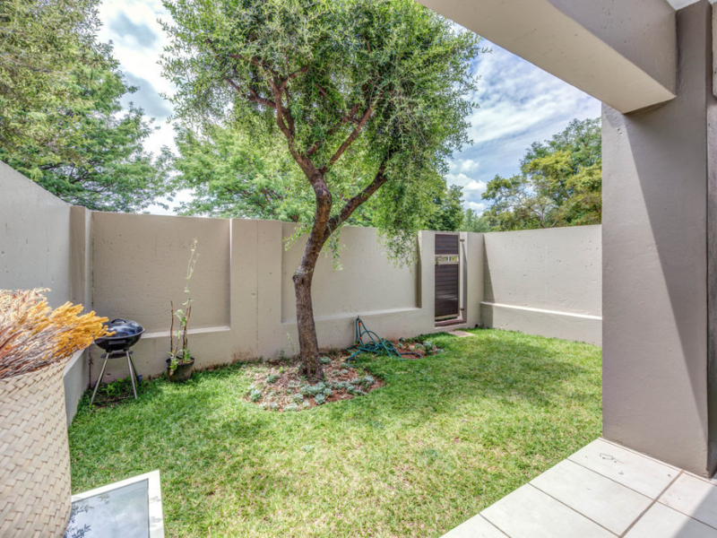 A private rear garden featuring a manicured lawn, a mature tree, and a paved patio area enclosed by high boundary walls.