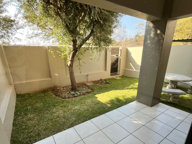 A covered patio area featuring tiled flooring and a concrete support pillar, overlooking a private garden space with a boundary wall.