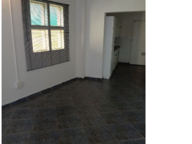 An empty living room featuring dark tiled flooring and a window with fitted venetian blinds, leading into an adjacent kitchen area.