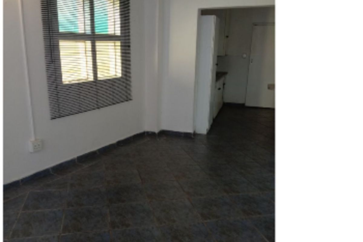 An empty living room featuring dark tiled flooring and a window with fitted venetian blinds, leading into an adjacent kitchen area.