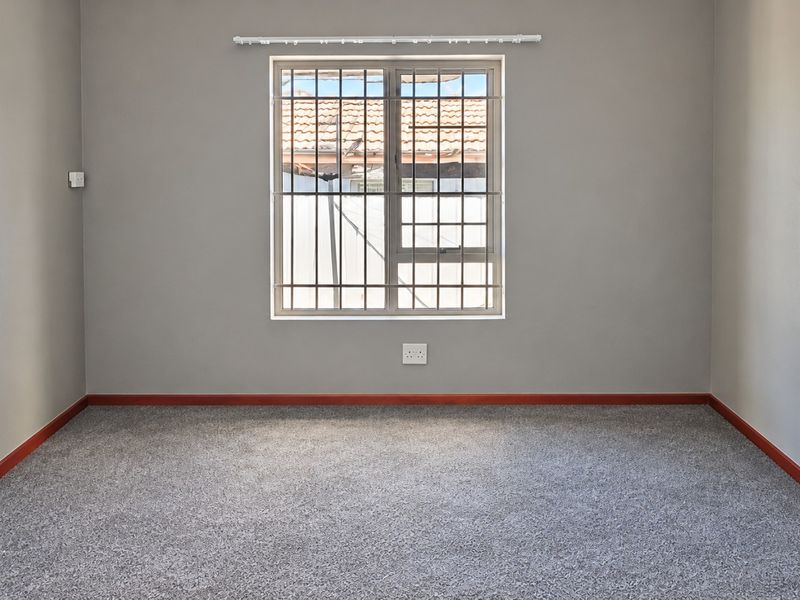 A clean, empty bedroom featuring neutral grey walls, carpeted flooring, and a window fitted with security bars.
