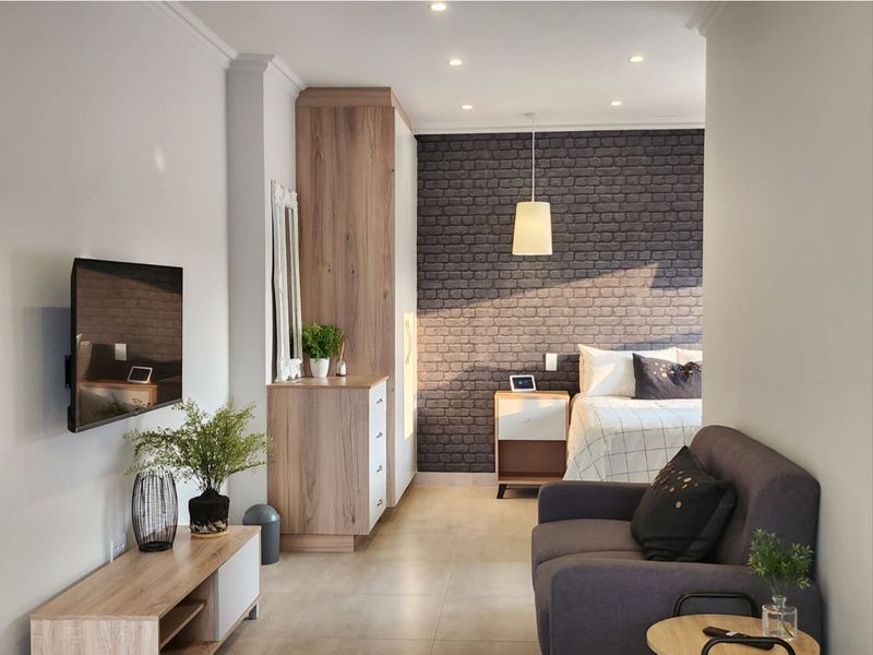 A modern open-plan studio apartment featuring a bedroom area with a brick-style accent wall, built-in cabinetry, and tiled flooring throughout.