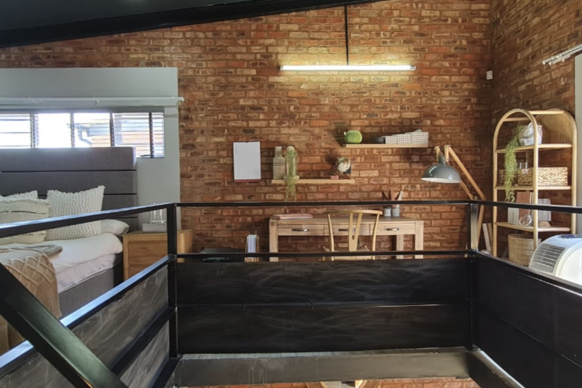A mezzanine bedroom featuring an exposed brick feature wall, industrial-style metal railing, and a sloped ceiling with integrated lighting.