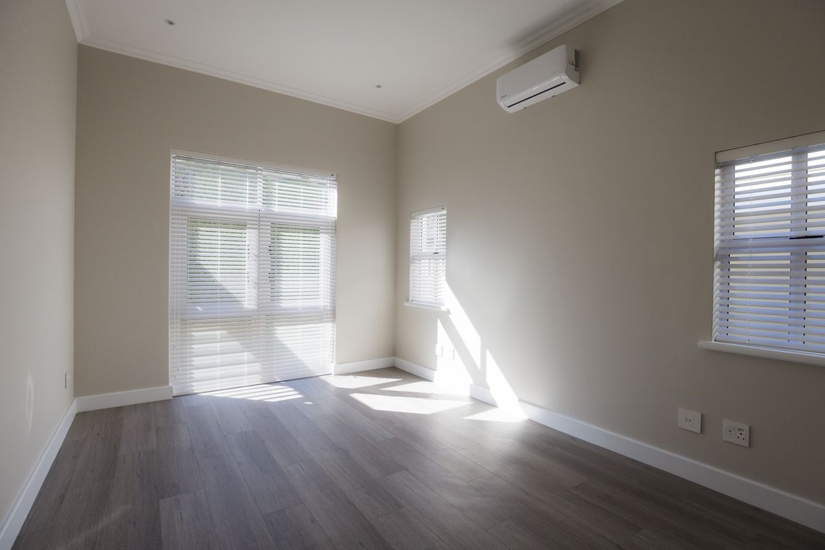 A bright, empty bedroom featuring modern laminate flooring, neutral walls, split-system air conditioning, and fitted venetian blinds.