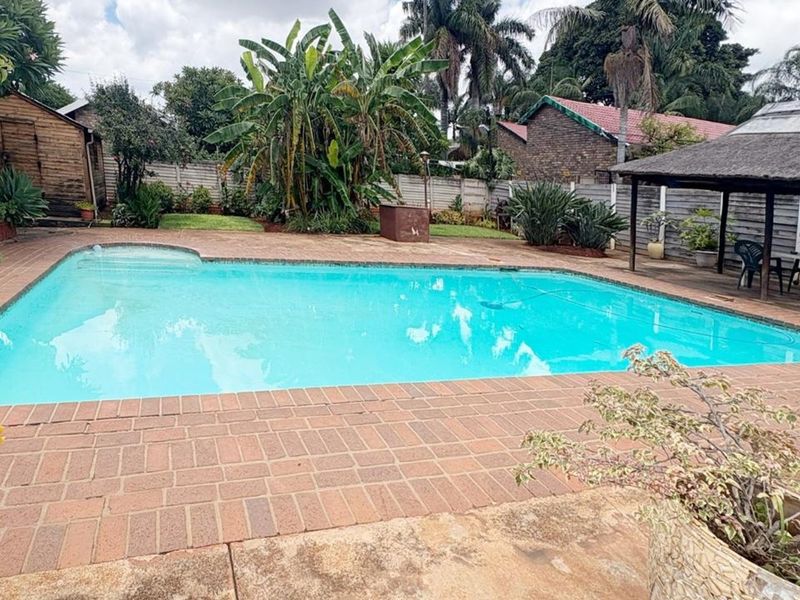 Outdoor pool area featuring a swimming pool, brick paved surround, and a thatched lapa, set within a landscaped garden with boundary walls.