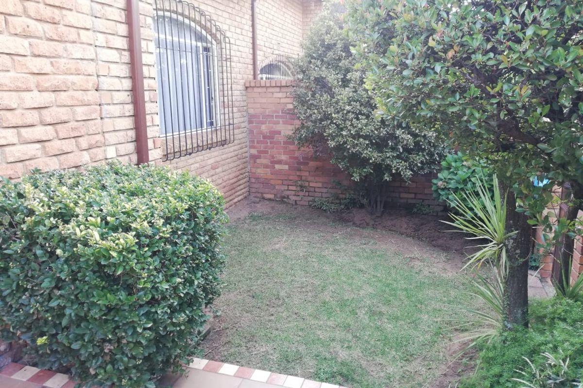 A private rear garden area featuring a face brick exterior wall, security-barred windows, and a small lawn bordered by established shrubs and trees.