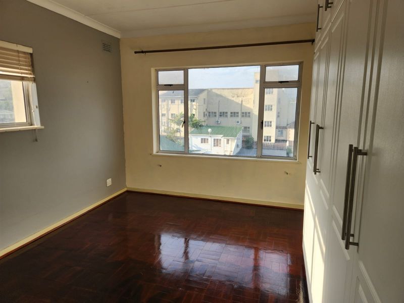 A bright bedroom featuring polished parquet flooring, built-in white cabinetry, and large windows providing natural light and an urban view.