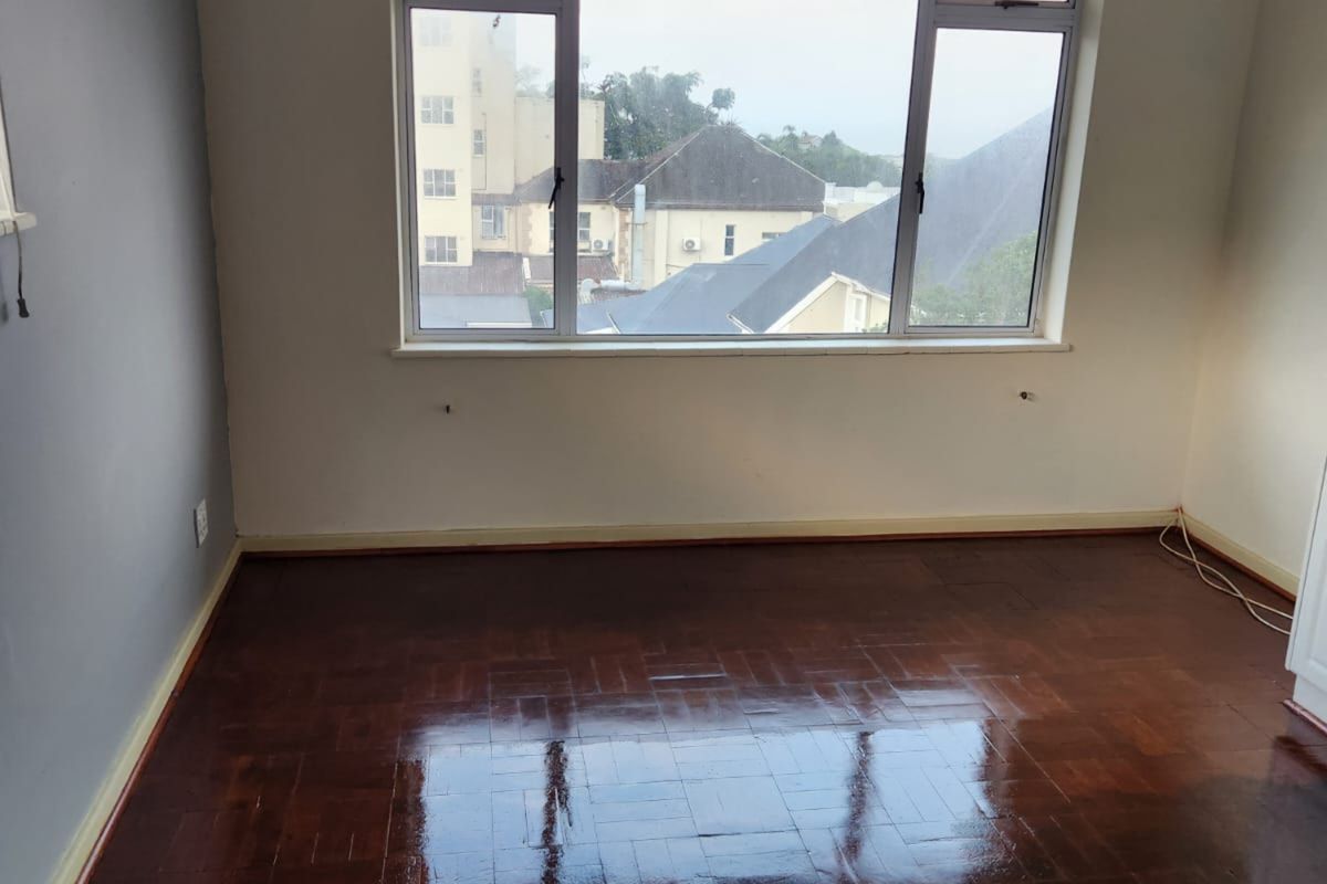 An empty bedroom featuring polished parquet wooden flooring, a large window with neighborhood views, and neutral wall finishes.