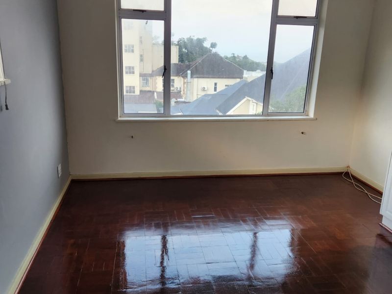 An empty bedroom featuring polished parquet wooden flooring, a large window with neighborhood views, and neutral wall finishes.