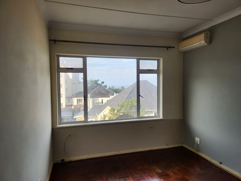 A vacant bedroom featuring polished parquet flooring, a large window with neighborhood views, and a wall-mounted air conditioning unit.