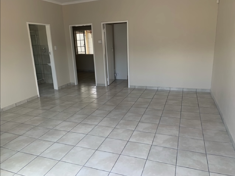 An empty living area featuring neutral tiled flooring and access to adjacent rooms, including a bathroom and hallway.