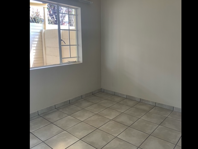 A bright, empty bedroom featuring neutral wall paint, tiled flooring, and a window with security bars overlooking the exterior area.