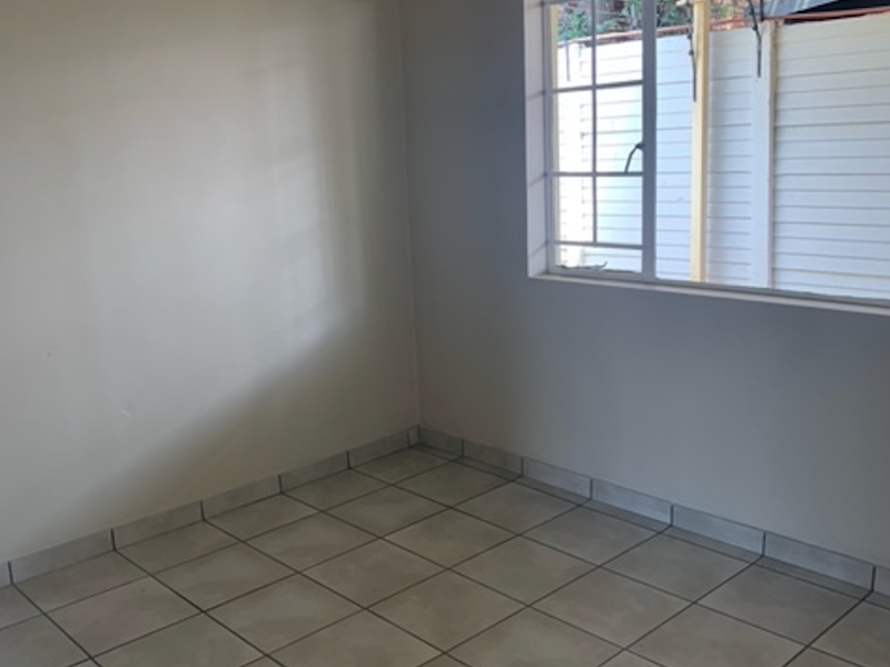 A bright, empty bedroom featuring neutral tiled flooring, plain walls, and a window with security bars overlooking the exterior area.