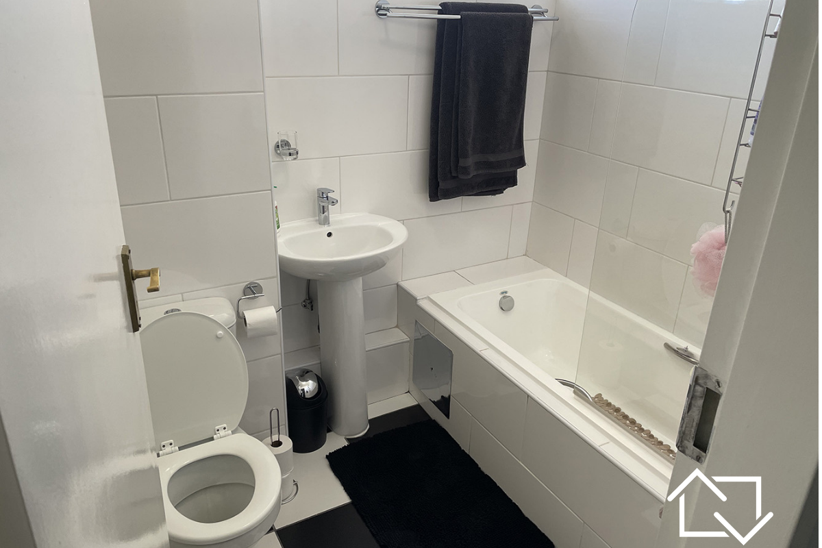A clean, functional bathroom featuring a pedestal basin, toilet, and a bathtub with a glass shower screen, finished with white wall tiles.